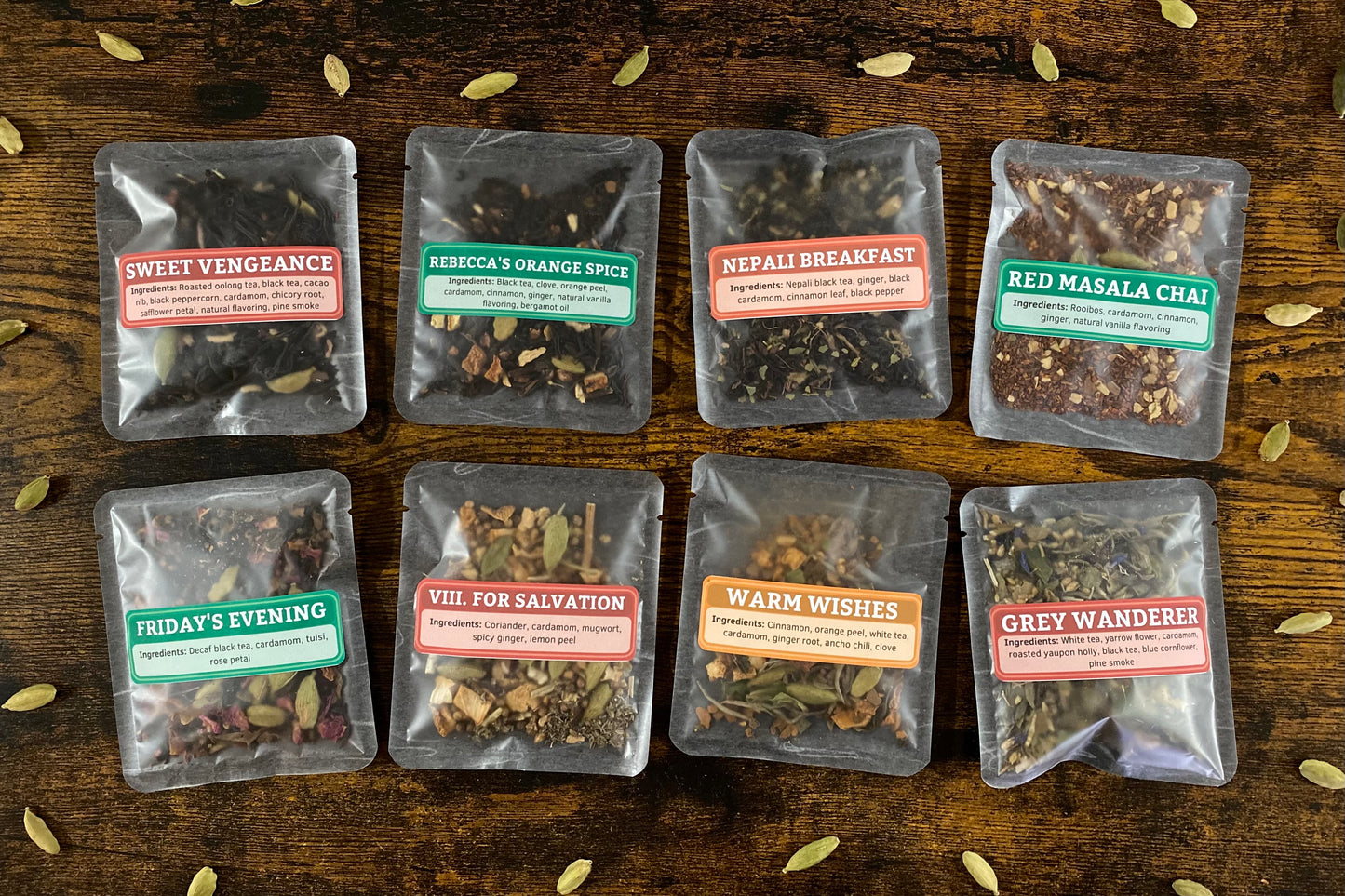 Eight packets of loose leaf chai samples on a wooden table surrounded by cardamom pods