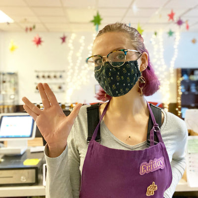 A brightly-lit tea house with a pink-haired woman wearing an apron with the nametag "Friday" holds her hands in a Vulcan peace salute.