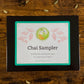 A square black box labeled 'Chai Sampler' on a rustic wooden surface scattered with cardamom seeds. 