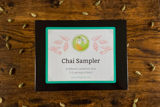 A square black box labeled 'Chai Sampler' on a rustic wooden surface scattered with cardamom seeds. 