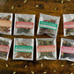 Eight individual tea bags arranged in two rows on a rustic wooden surface. Each clear resealable pouch has a colorful label naming a different tea variety: Cocoa Royal Tea, Dungeon Cardamom Carl, Friday's Evening, The Chai of the Bedlam Bride, Grey Wanderer, Red Masala Chai, Nepali Breakfast, and Harvest Spice.