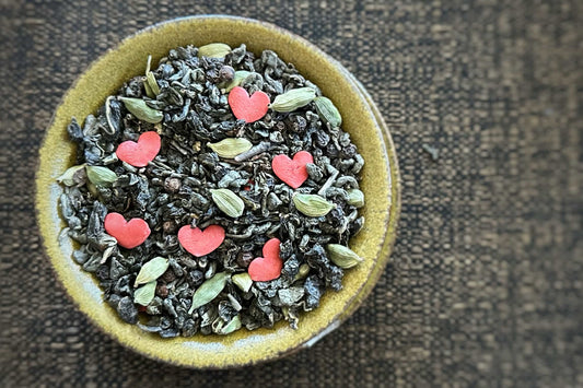 Green teacup full of rolled green and oolong tea with cardamom pods, black peppercorn and red heart sprinkles