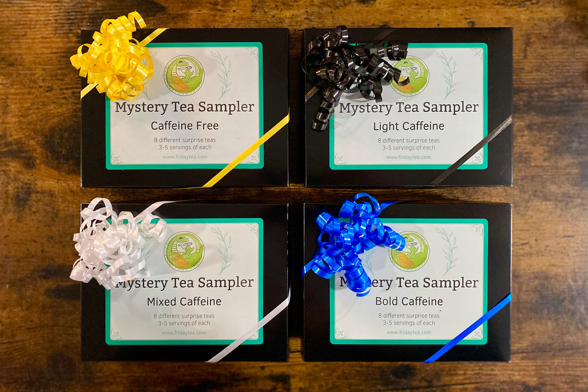 Four tea sampler boxes arranged in a 2x2 grid on a wooden surface, each with a ribbon bow: yellow, black, white, and blue. The labels read 'Mystery Tea Sampler' with caffeine level options 'Caffeine Free,' 'Light Caffeine,' 'Mixed Caffeine,' and 'Bold Caffeine.