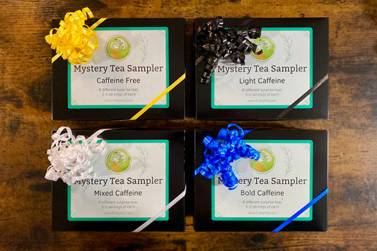 Four tea sampler boxes arranged in a 2x2 grid on a wooden surface, each with a ribbon bow: yellow, black, white, and blue. The labels read 'Mystery Tea Sampler' with caffeine level options 'Caffeine Free,' 'Light Caffeine,' 'Mixed Caffeine,' and 'Bold Caffeine.